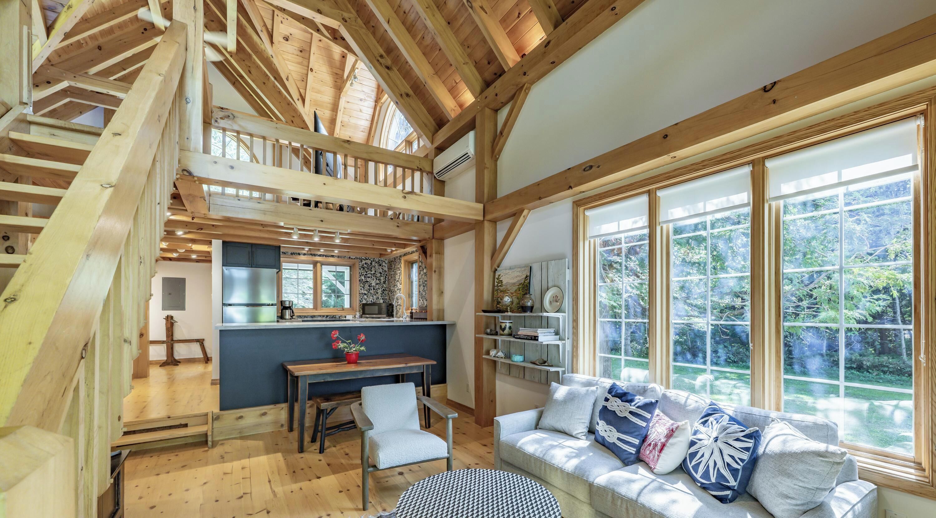 A spacious loft-style room with a high ceiling, wooden beams, a dining area, a sofa with decorative pillows, and large windows offering a view of the outdoors.
