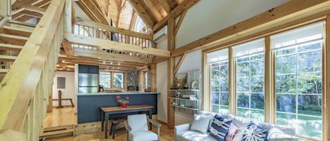 A spacious loft-style room with a high ceiling, wooden beams, a dining area, a sofa with decorative pillows, and large windows offering a view of the outdoors.