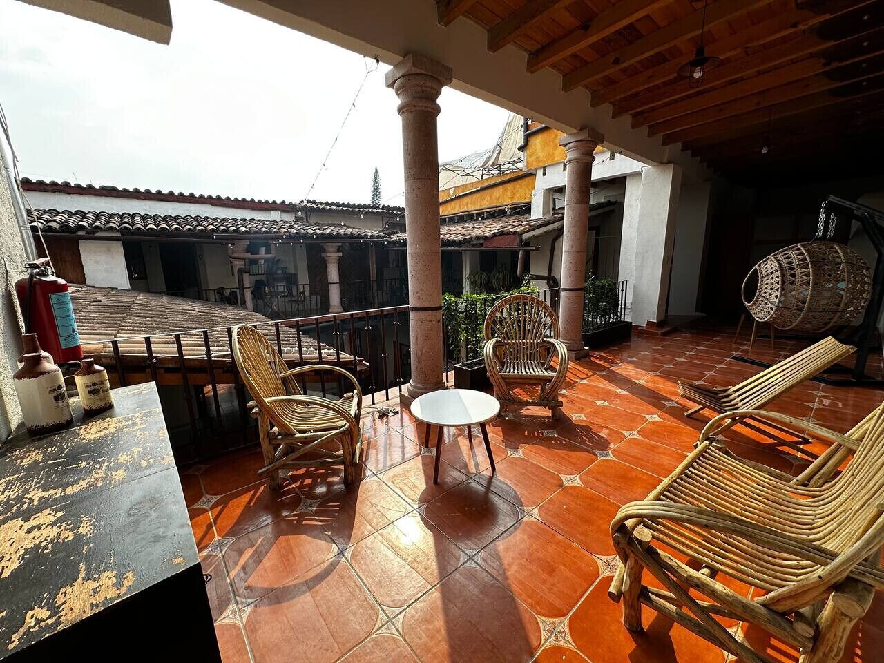 A covered outdoor seating area with wicker chairs and a table, surrounded by tiled flooring and columns.