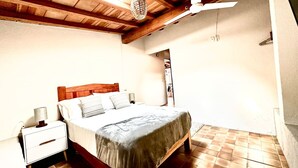 Un dormitorio con techo de madera, una cama con cubrecama gris, una mesita de noche con lámpara y suelo de baldosas.