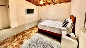Un dormitorio con techo de madera, una cama con colchón blanco y almohadas a rayas, una mesita de noche con lámpara y un televisor montado en la pared.