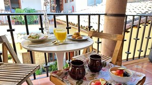 Une table dressée pour un repas, avec un saladier de fruits, deux tasses et un verre de jus d’orange, sur un balcon offrant une vue sur des bâtiments et de la verdure.