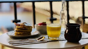 Une table avec une pile de crêpes, un verre de jus d’orange et une carafe sombre sur un balcon.