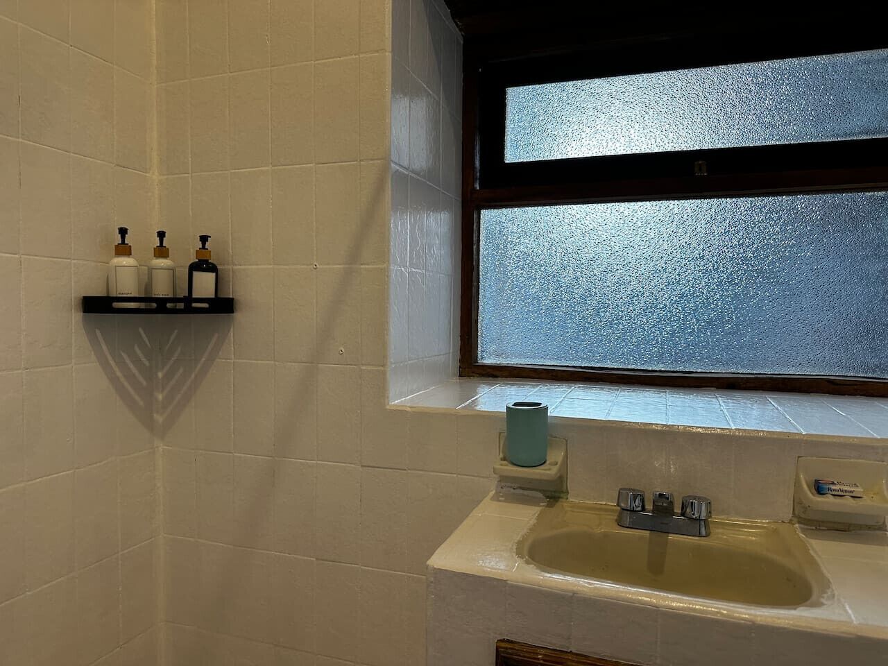 Ein Badezimmer mit weiß gefliesten Wänden, einem Fenster, einem Waschbecken mit Wasserhahn und einem Ablagebereich mit Pflegeprodukten.