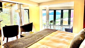 Een hotelkamer met een bed, twee stoelen, een tafel en uitzicht op de zee door grote ramen.