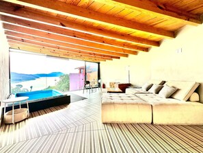A modern interior with a large window overlooking a pool and mountains, a sofa, and a wooden ceiling.