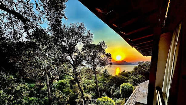 A view from a balcony overlooking a lush garden and a sunset over a body of water. - Vista Rio Bosque Apartment, Heated Pool 6 (Valle de Bravo)
