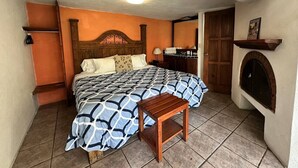 A bedroom with a bed, wooden headboard, bedside tables, a nightstand, a small wooden stool, a fireplace, and a door. - Vista Rio Bosque Apartment, Heated Pool 6 (Valle de Bravo)