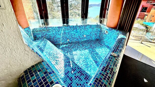 A glass-enclosed hot tub with a blue mosaic tile interior, surrounded by a balcony with a view of trees and buildings. - Jacuzzi Apartment Lake View, Heated Pool 7 (Valle de Bravo)