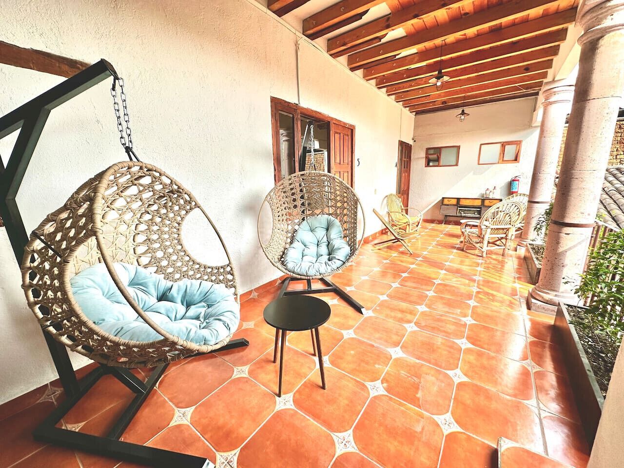 A covered patio with wicker chairs and a small table, tiled flooring, and wooden beams.
