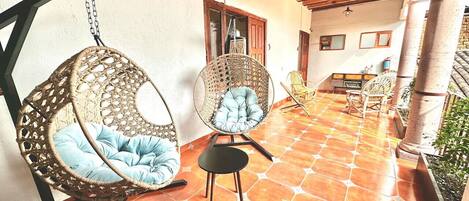 A covered patio with wicker chairs and a small table, tiled flooring, and wooden beams.