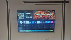 A flat-screen TV mounted on a wall displaying a food-related TikTok advertisement. Below the TV, there is a white entertainment unit with a vase of flowers and various items. - Modern vacation apartment in a very beautiful location, surrounded by forests and hiking trails! (Heimbuchenthal)