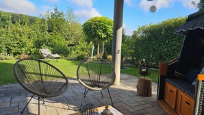 An outdoor patio with two wicker chairs, a wooden table with a vase, and a grill. - Modern vacation apartment in a very beautiful location, surrounded by forests and hiking trails! (Heimbuchenthal)