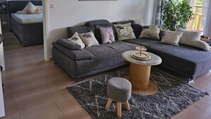 A modern living room with a grey sectional sofa, a round coffee table, and a grey rug. There is a painting on the wall and a plant by the window. - Modern vacation apartment in a very beautiful location, surrounded by forests and hiking trails! (Heimbuchenthal)