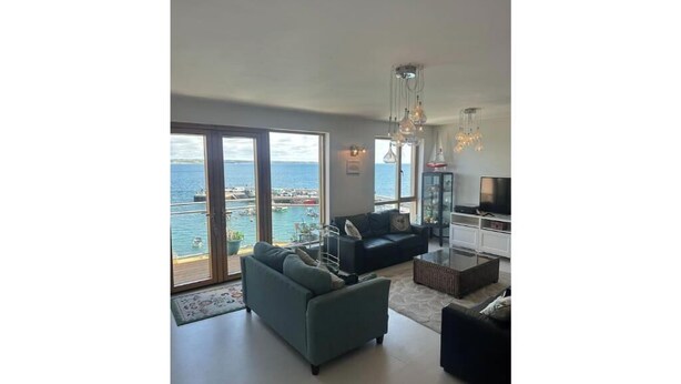 A modern living room with a large sliding glass door leading to a balcony with a view of the sea. There is a sofa, a coffee table, and a TV stand. - Atlantic Views & Irish Charm (Ballycotton)