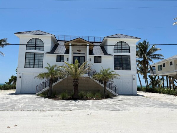 A modern two-story house with a central entrance, a covered staircase, and a view of the beach.