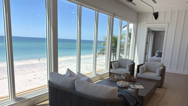 A spacious living room with large windows overlooking a beach, a grey sofa, a coffee table, and a chair.