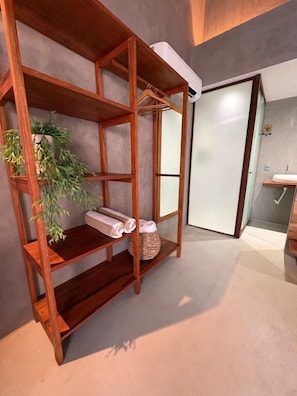 A wooden shelf unit with towels and a plant, a bathroom with a sink and mirror, and a door leading to another room.
