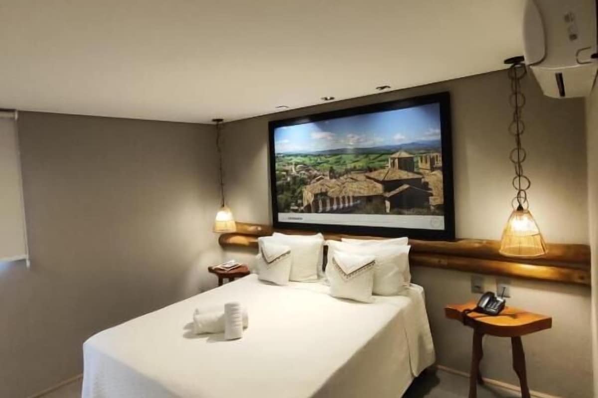 A hotel room with a bed, a wall-mounted TV, a bedside table, and a lamp.