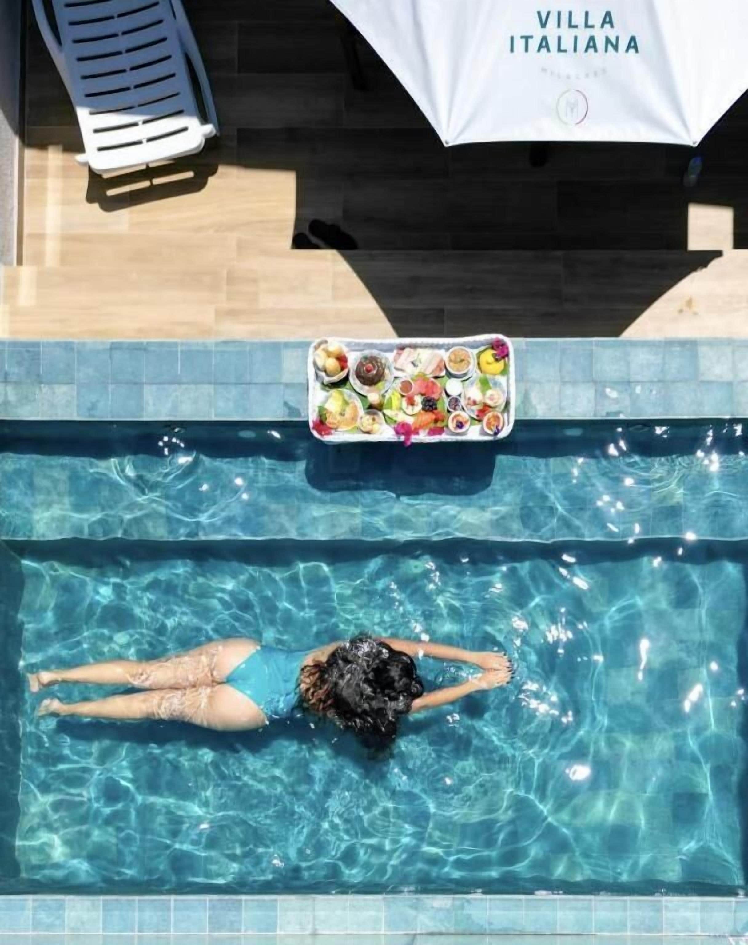 Una piscina con una persona flotando, una silla de descanso y una mesa con comida.