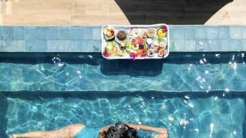 A swimming pool with a person floating, a lounge chair, and a table with food items.