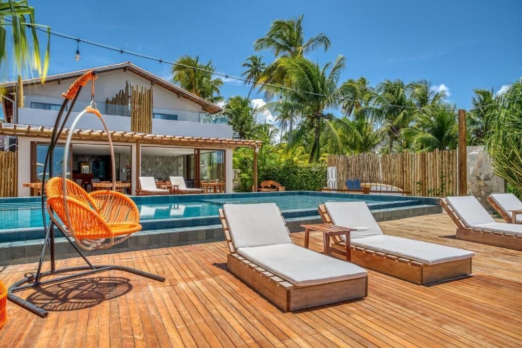 Un área junto a la piscina con sillas de descanso y una hamaca colgante, rodeada por una terraza de madera y palmeras.
