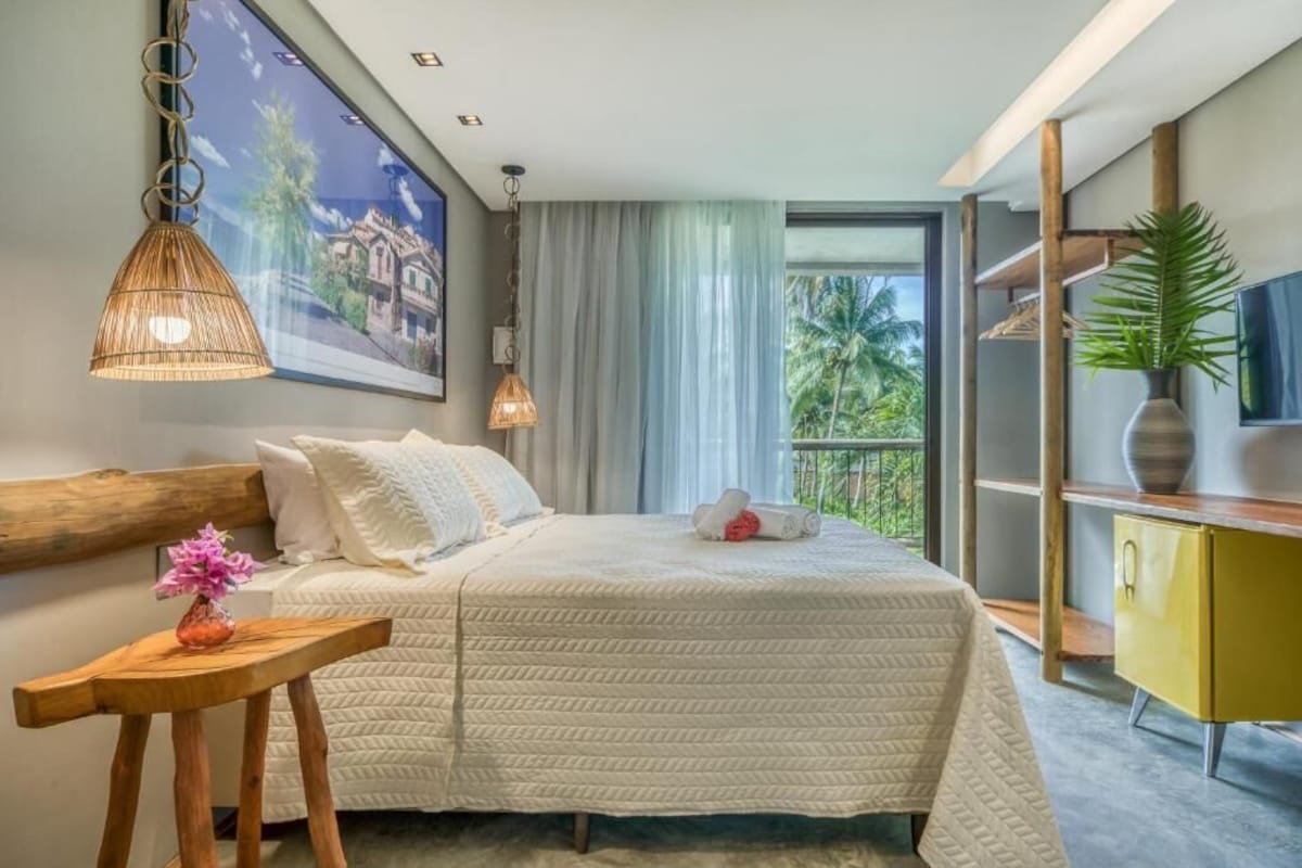 A hotel room with a large bed, a wooden headboard, a small table with a vase, a television, and a balcony with a view of palm trees.