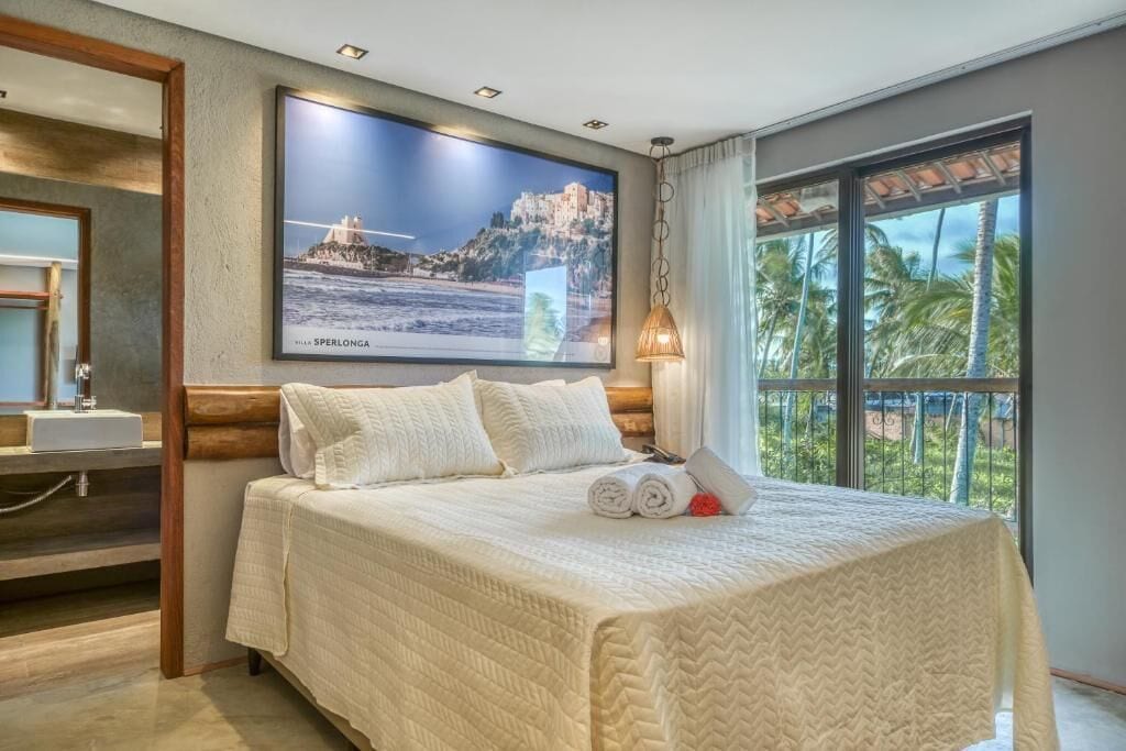 Quarto de hotel com uma cama grande, televisão na parede, janela com vista para palmeiras e cabeceira de madeira.