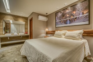 A hotel room with a large bed, a wooden headboard, a large mirror, a wall-mounted air conditioner, and a painting above the bed. - Villa Italiana (São Miguel dos Milagres)