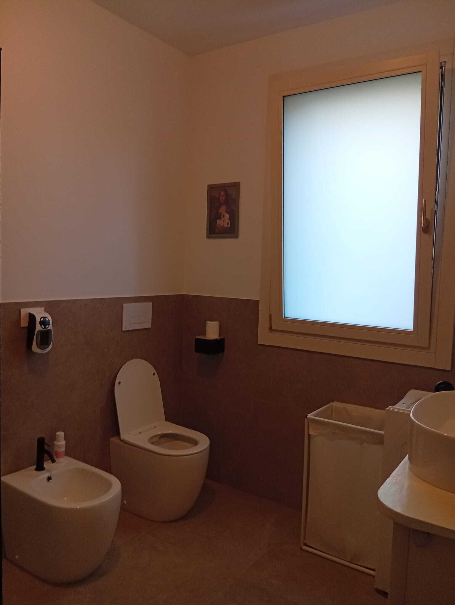 A bathroom with a toilet, bidet, and sink. There is a window with blinds, a small picture on the wall, and a tissue dispenser.