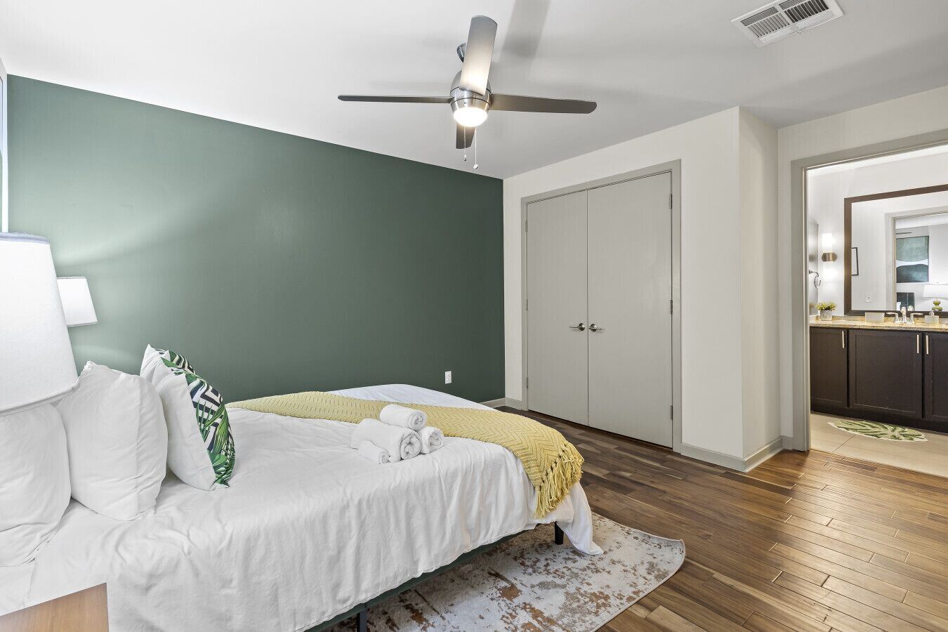 Une chambre à coucher avec un lit, un ventilateur de plafond, une armoire et une salle de bain visible à travers une porte ouverte.