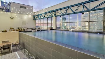 A rooftop swimming pool with a view of the city skyline, a wooden deck, and a covered area with seating.