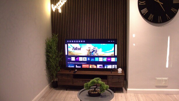 A modern living room with a flat-screen TV displaying 'Fallout', a potted plant, a bonsai tree, and a large wall clock. - Luxury 3 bedroom house in Harlow, Essex (Harlow)