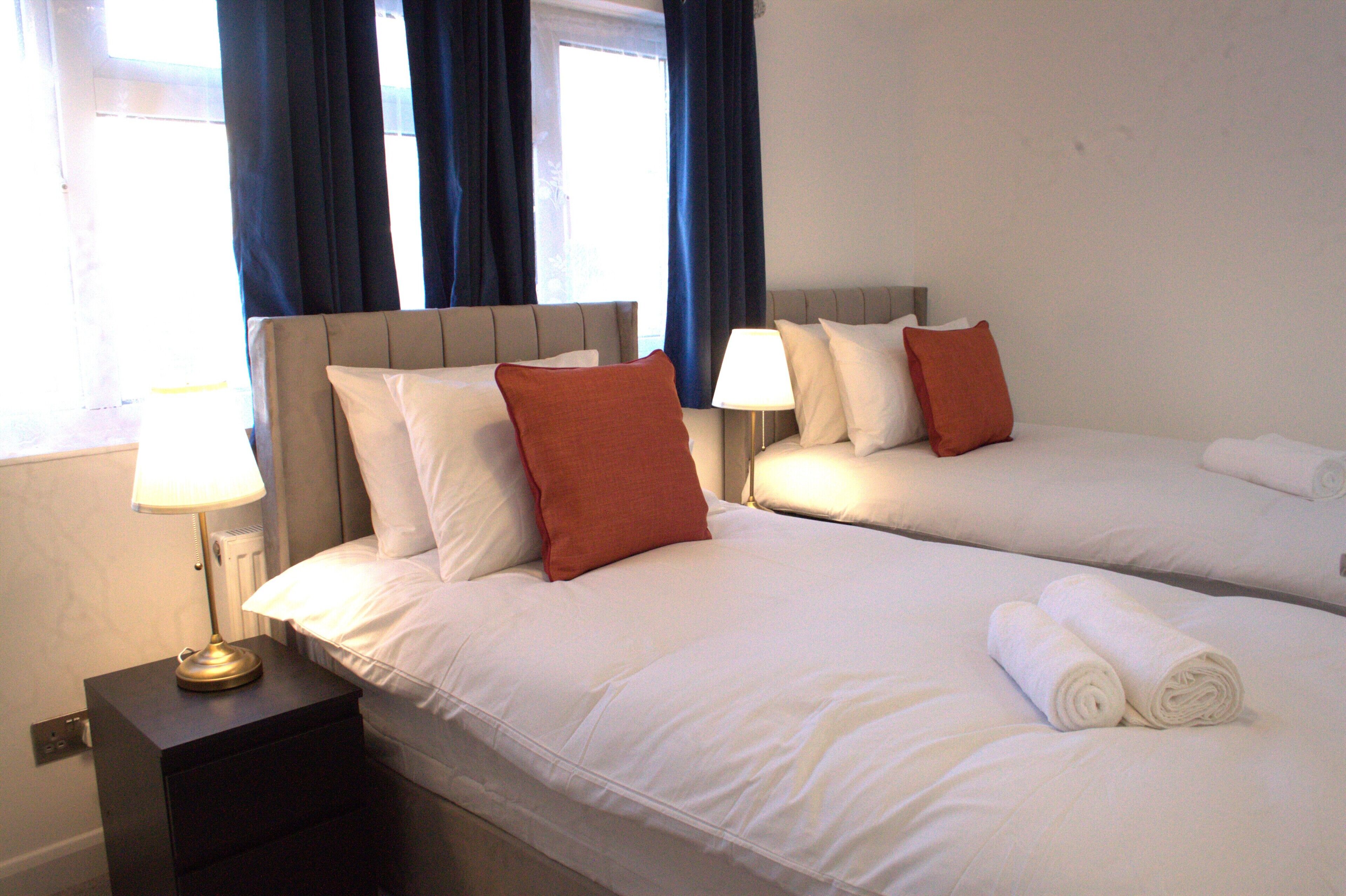 A hotel room with two beds, each with white bedding and red pillows, a bedside table with a lamp, and a window with blue curtains.