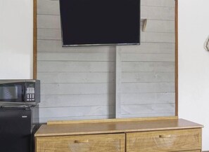 Quarto com televisão de tela plana fixada em parede de painel de madeira, micro-ondas sobre um pequeno eletrodoméstico e armário de madeira com gavetas.