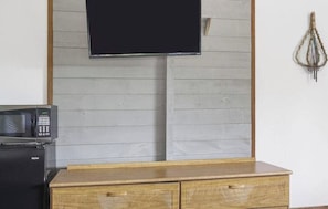 Una habitación con una televisión de pantalla plana montada en una pared de paneles de madera, un microondas sobre un mueble de madera y un adorno colgando en la pared.