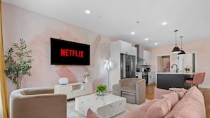 A modern living room with a large flat-screen TV, a sofa, a coffee table, and a dining area with a kitchen in the background.