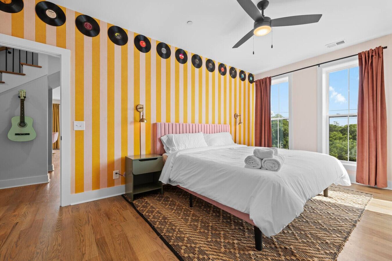 Une chambre avec un mur à rayures, un lit avec une literie blanche, une guitare et un ventilateur de plafond.