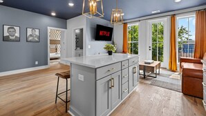 A modern kitchen with grey cabinetry, a central island, pendant lighting, and a large window with curtains. - NASH Bach Pad 3-Bedroom Penthouse near Downtown Nashville (Sleeps 12) (Nashville)