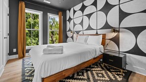 A modern bedroom with a large bed, a nightstand, a wall with a geometric pattern, and a view of greenery through the windows. - NASH Bach Pad 3-Bedroom Penthouse near Downtown Nashville (Sleeps 12) (Nashville)