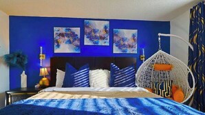 Una camera da letto con un letto, decorazioni murali blu, una poltrona sospesa e un comodino con una lampada.