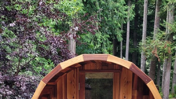A wooden barrel-shaped structure with a bench inside, set on a concrete base in a forested area.