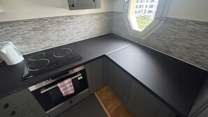 A compact kitchen with a black countertop, grey cabinets, a built-in oven, and a window with a view of buildings.