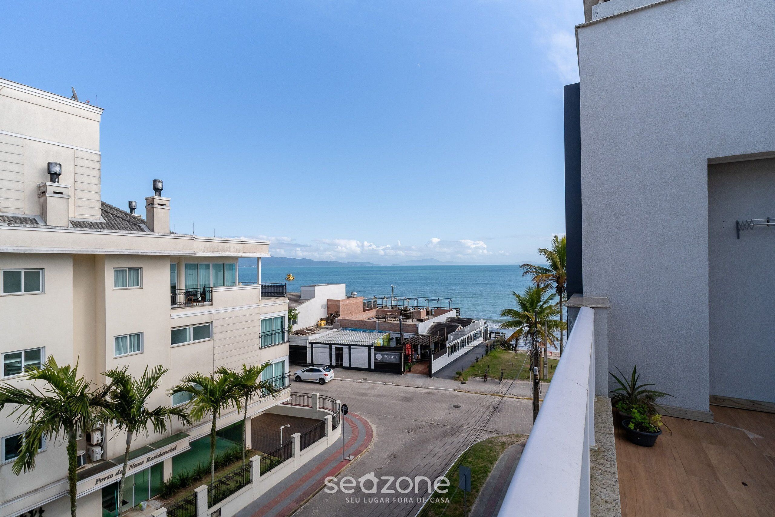 A residential building with a view of the sea, a parking area, and a few palm trees.