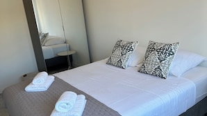 A neatly made bed with white linens and two patterned pillows, a grey quilted bedspread, and two rolled towels on the bed. - Right in the town center, 5m from the beach on foot and 2m from the SNCF train station (Saint-Raphaël)