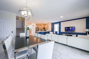 A modern living room with a dining table, white sofas, a TV, and a kitchen in the background.