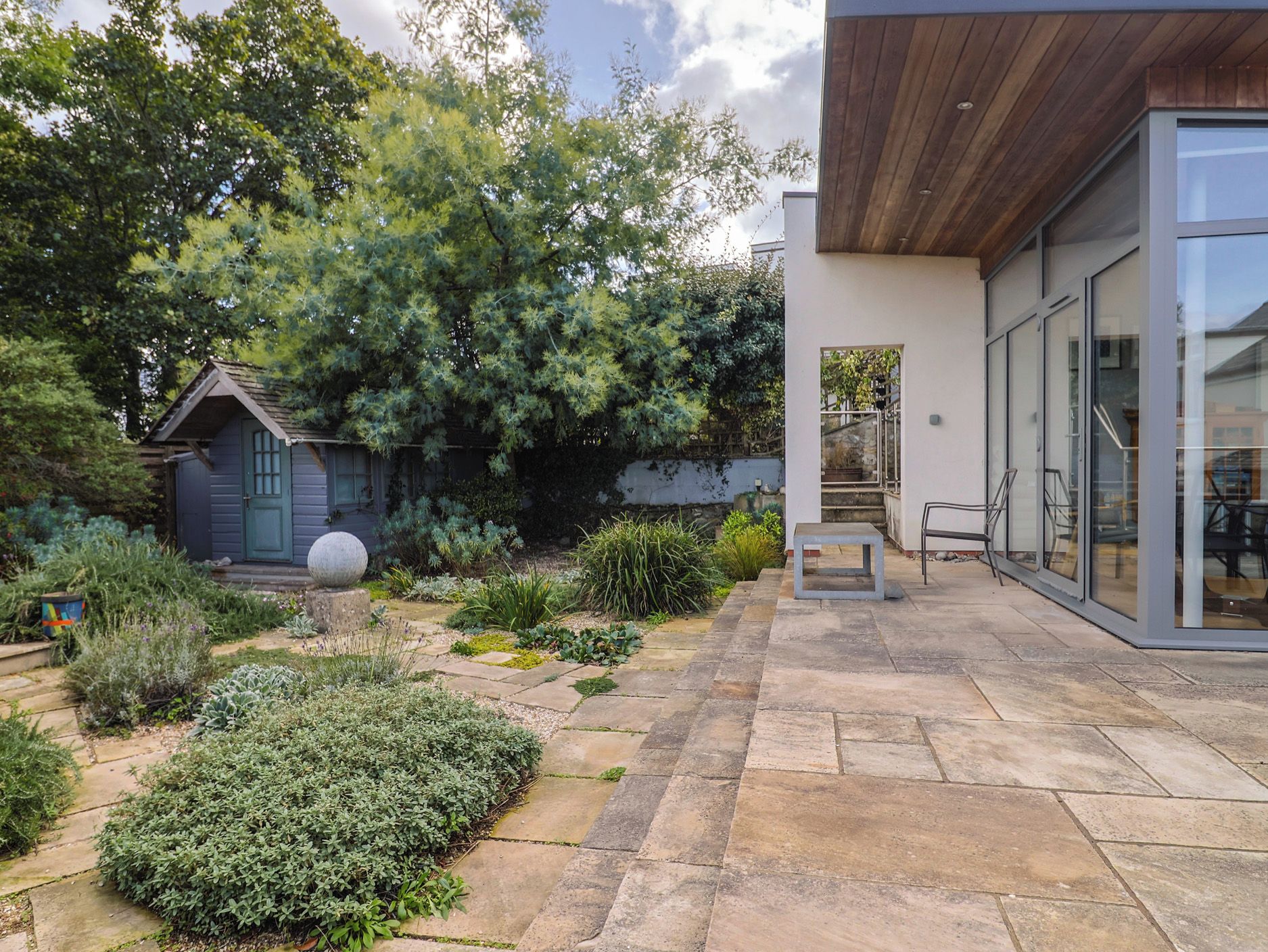 Una casa moderna con un patio de piedra, un cobertizo pequeño y una variedad de plantas.