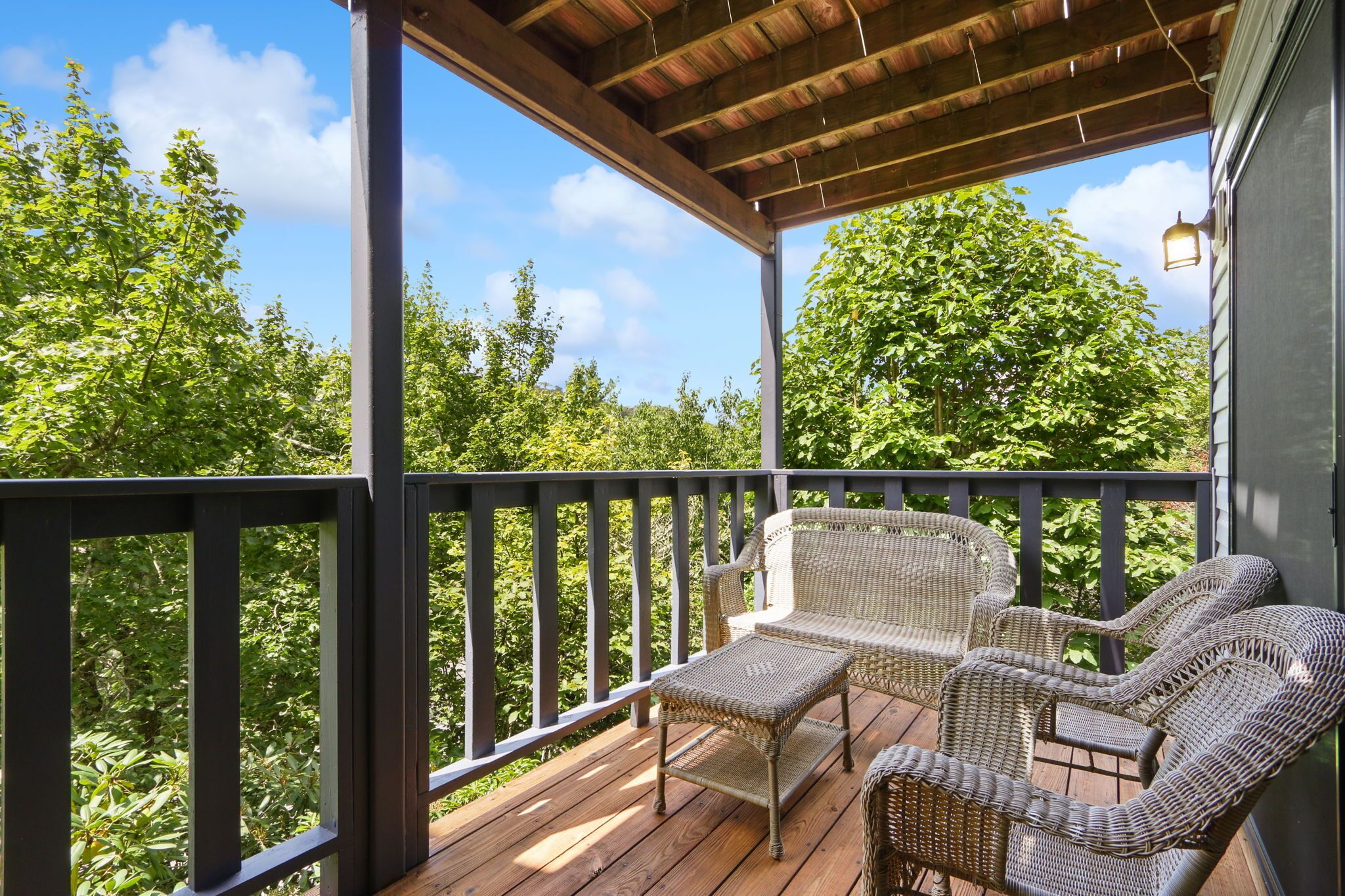 A covered outdoor deck with wicker furniture and a railing, overlooking a lush green landscape.