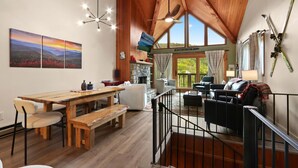A spacious living area with a wooden ceiling, a dining table with benches, a fireplace, and a balcony with ski equipment.