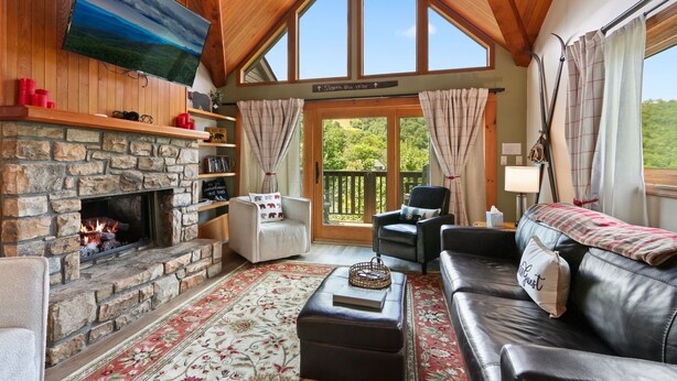 A cozy living room with a stone fireplace, a flat-screen TV, a comfortable sofa, and a view of the outdoors through large windows.